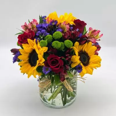 Reminiscent of strolling through a field of flowers, this colorful arrangement captures features magnificent sunflowers, alstroemeria and poms set in a trendy mason jar with a natural raffia bow.