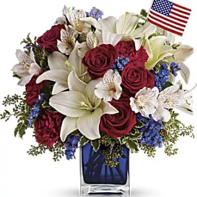 A patriotic pick to send a brave veteran, decorate your Fourth of July picnic, or celebrate Memorial Day. Lush red, white and blue flowers are presented in a deep blue glass cube vase along with an American flag.
Red roses, white asiatic lilies, white alstroemeria, blue statice and red carnations are gathered with green pitta negra, seeded eucalyptus and a small American flag in a cobalt blue glass cube.